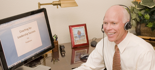 Dr. Merlin Switzer using headphones in front of the computer providing coaching over the phone with the banner, Coaching for Leadership Success, written on the screen.